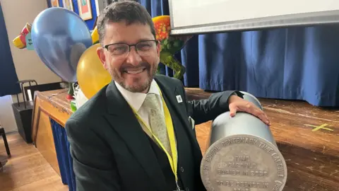 John Devine/BBC Craig Jansen has short brown hair and is wearing black framed glasses, a white shirt, black suit jacket and green tie. He is holding a silver metal time capsule to the right.