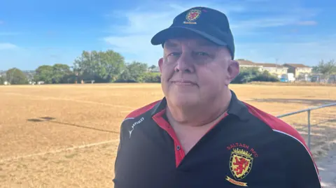Nick Rich from Saltash Rugby Football Club stood wearing a black and red rugby polo shirt and black branded hat. He is looking at the camera with a dry yellow coloured pitch behind him.