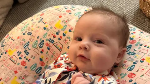 Reagan Thomas A baby with a small amount of brown hair wearing a floral patterned onesie lying on a pink cushion.