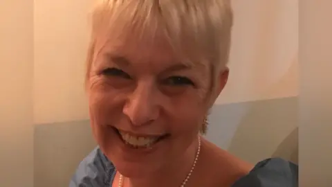 A woman with short, light-coloured hair wearing a blue top. A pearl necklace is visible around the neck. The background consists of a plain wall.