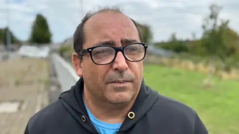 Michael O'Brien. He wears glasses and is bald, with dark short hair on the sides. He has short stubble and wears a black hoodie, with the background blurred behind him. 