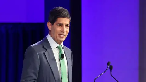 Donald Trump's Fed pick Kevin Warsh addresses an audience from a lectern, wearing a navy suit with a green tie. 