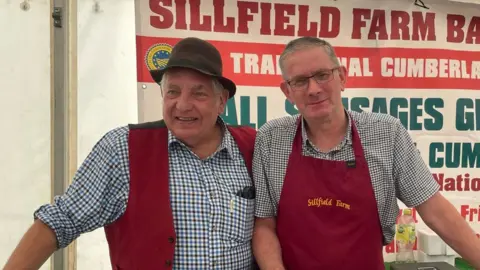 Peter Gott and a colleague standing in a market stall as they sell sausages. They are dressed in chequered shirts and red aprons. Peter is wearing a brown hat.