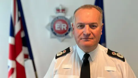 Chief Constable Stephen Watson is starring directly at the camera in his police uniform. The Greater Manchester Police logo is hanging on the wall behind him and there are Union Jack flags on either side.
