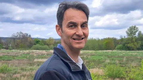 PA Tim Slaney, a white man in his sixties with short greying dark hair looks at the camera smiling with South Downs National Park greenery in the background
