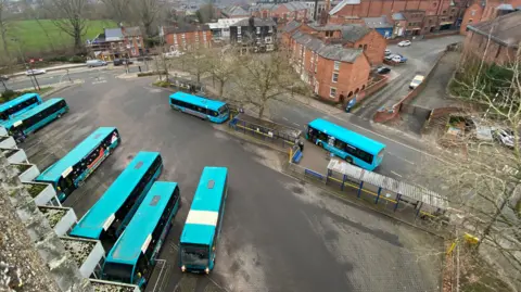 Eight turquoise single-decker buses parked in a large bus station. Houses and a green field can be seen in the background.