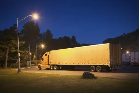 A white lorry is parked up for the night at a truck stop under a  street lamp. In the background you can see two other HGV's and around them is tree's and grass.