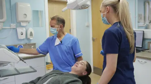 Glasgow dentist David McColl with a patient undergoing a check-up - she is lying down and smiling. He is in his medical scrubs and checking a machine. A dental nurse is standing next to the patient, with her back to the camera.   