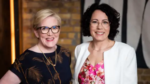 With blonde hair and wearing a black blouse Monika Liikamaa stands next to Denise Johansson who wears a white jacket and flowery pink blouse.