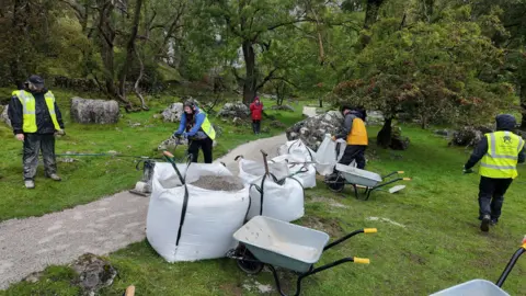 Yorkshire Dales National Park Authority Several people in high‑visibility vests and rain gear working on a woodland path, using wheelbarrows and shovels to spread material from large white bags, surrounded by grass, trees and scattered rocks.