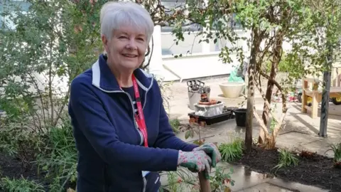 MCHT A woman with short white hair, a blue jacket and red lanyard smiles as she has both her hands on the handle of a spade, which she was putting into the ground. She is standing in the middle of a garden.