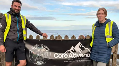 The Community Traverse David Keane and Kitty-Leigh Oxley stand by the sea at Robin Hood's Bay. They both stand with fluorescent jackets on and running gear. They stand in front of a black sign reading Core Adventures.