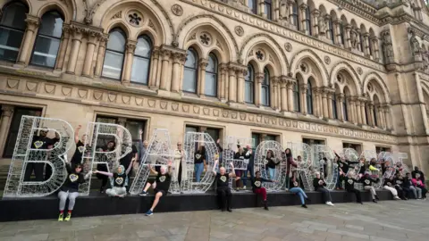 A group of people in black T shirts stand around 6-8ft letters that read 'Bradford' in front of Bradford City Hall. 