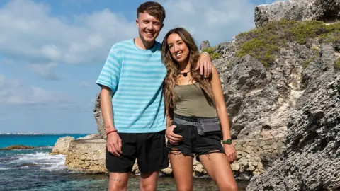 StudioLambert/BBC Roman Kemp has him arm around sister Harleymoon as they both smile on a path next to the sea next to a large rock.