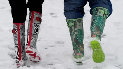 PA Media A close up from behind showing the legs of two youngsters walking in brightly-patterned wellies in the snow. The person on the left has black trousers tucked into zebra print and red wellies. The child on the right has boots with a green ad khaki swirling pattern.