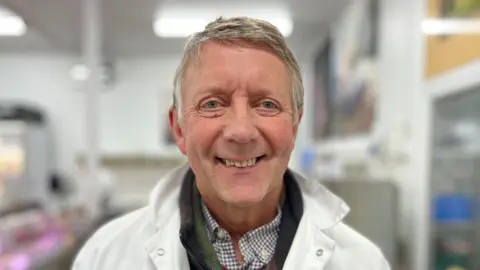 Martin Giles/BBC A man smiles at the camera. He has short grey hair and is wearing a white butchers jacket with a chequered shirt underneath and green jumper. The background is blurred.