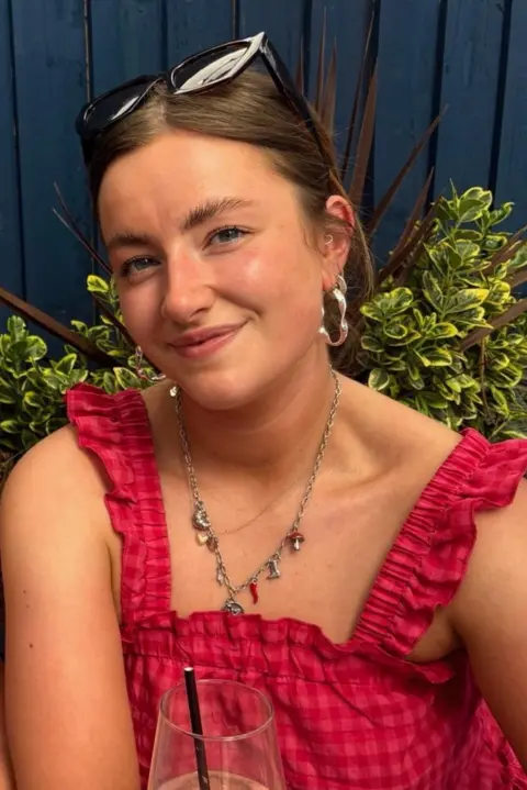 Erin Gardner Erin Gardener smiles at the camera. she is wearing a bright pink top and gold earrings. she has brown hair and there is some plants in the background.