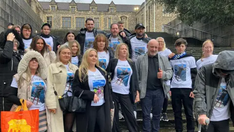 A group of 22 people standing on the steps of Winchester Crown Court. Nearly all of them are wearing white t-shirts printed with pictures of Joey Johnston which read Justice for Daddy and Justice for Joey.