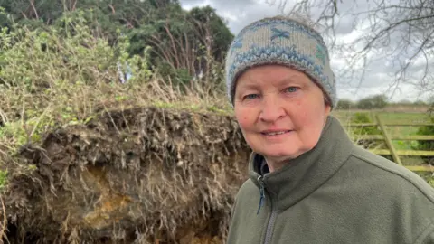 Emma Rawlings has a green jacket and is wearing a blue hat. She has blue eyes. She is standing in front of an uprooted tree.