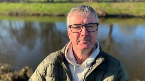 Photograph of angler Mike Duddy. He is pictured in front of the River Irwell in the Agecroft area of Salford.