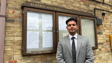 Martin Heath/BBC Tushar Kumar in a grey suit stands beside a noticeboard outside Elstree and Borehamwood Town Council, set against a light brick wall. The board is filled with official printed documents pinned neatly in rows. The formal setting and smart attire give the image a composed, civic feel.