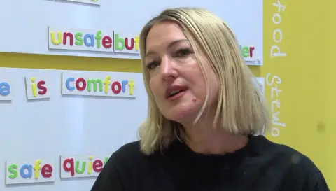 Lucy Groenewoud has short blonde hair, dark eyes and is wearing a black t-shirt, in the background is a colourful board of words to help people feel accepted.