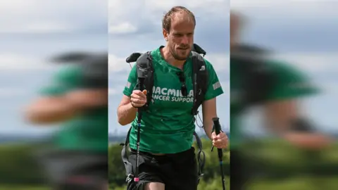 PA Media Mr Dye wearing a green T-shirt that reads: Macmillan Cancer Support, carrying walking poles and wearing a black rucksack. He is walking in the countryside.