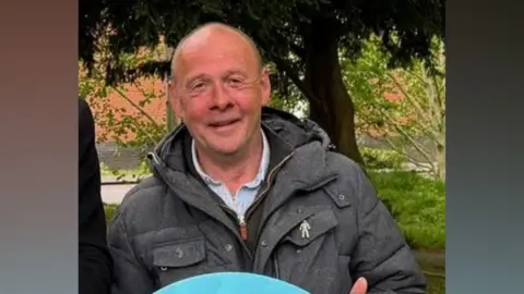 Tierney is stood outside by some trees and is smiling at the camera. He has a grey jacket on over a grey shirt. He has short brown hair.