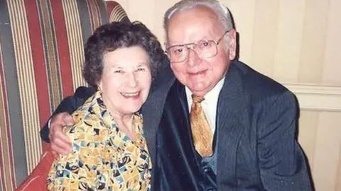 Family handout Una Crown, a dark grey-haired woman is wearing a yellow-patterned dress sitting sitting next to her husband, a grey haired man, who is wearing a blue jacket and blue waistcoat and yellow tie. He has his right arm around the woman's shoulders.