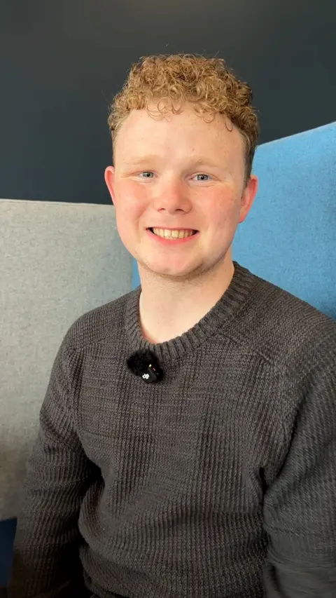 Man sitting down in grey jumper smiles at camera with light blue and grey background.
