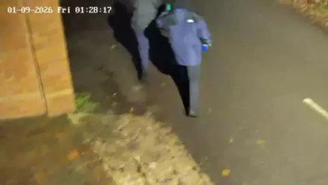 Camran Butt CCTV footage showing two men, wearing gloves and with their hoods raised, walking away from the camera. One is carrying a blue bottle in his right hand. 