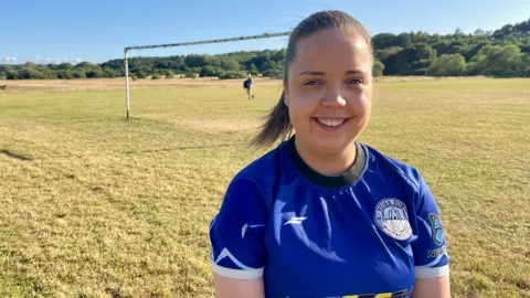 A woman in blue football shirt stands on a grass roots football pitch. She has dark hair tied in a ponytail. Her shirt displays the club badge of Stoke Town Football Club and a clothing logo. Rusty goal posts and trees can be seen in the distance.