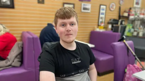 Liam has short ginger hair. He is sitting in a cafe with comfy-looking purple seats and is wearing a black t-shirt.