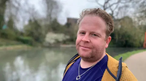 Ben Jelley is wearing his wife's engagement and wedding rings on a necklace. He is standing beside the canal and is wearing a blue t-shirt and yellow jacket.