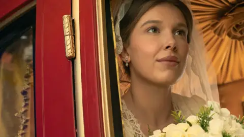 Netflix Millie Bobby Brown as Enola Holmes wearing a wedding dress and holding flowers 