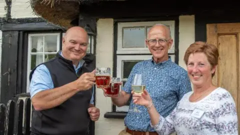 Dave Denny Dave stands between two other volunteers - a male on the left and female on the right - outside the pub, chinking glasses of beer and white wine together in celebration. They are all smiling to camera. They are under the eaves of the pub, with a window right behind them and part of a wooden door to the right