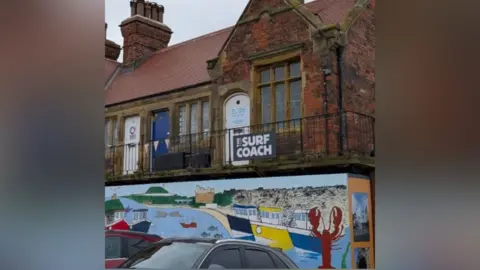 North Yorkshire Council An elevated building with railings around the front which has a banner attached which says 'Surf Coach'. There is boarding around the lower half with colourful drawing depicting a beach scene. 