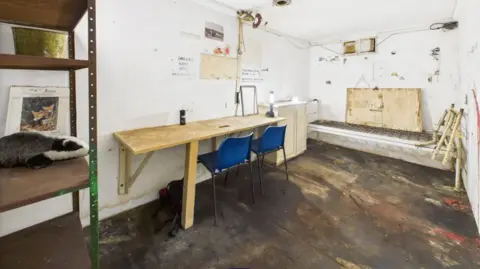 Interior of the bunker which has white walls a small wooden desk and two blue plastic chairs. At the near left side, there is a basic-looking stack of shelves which are brown and stained.