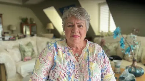 A woman with a shorter, grey hair wears a colourful overshirt, featuring blue, green, yellow and pink flowers of different sizes over a white base. She has a necklace with a green pendant. She's sat in her living room, and in the background are two grey sofas, with a vase of vibrant blue flowers over her left shoulder.
