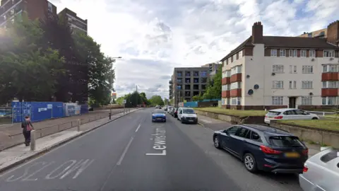 A Google street view screengrab of a road running through a street lined with parked cars, buildings and trees. A pedestrian walks along the left pavement.