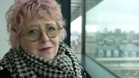 BBC Close-up of a middle-aged woman with short pink hair and round green glasses. She is wearing a black and white checked scarf and looks sorrowfully at the camera. 