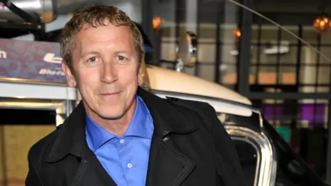 Getty Images A man wearing a blue collared shirt and a black jacket stands with his hands behind his back while looking at the camera.