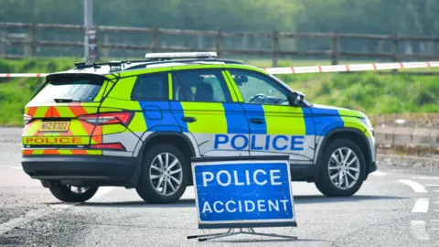 A blue, yellow and red vehicle with red police label on the doors and boot. The car is parked across a road blocking it and there is a blue sign in the foreground with the words Police accident in white writing. 
