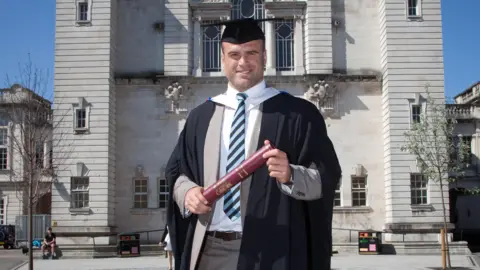 Cardiff University Jamie Roberts stands in front of a grey building while holding a scroll. He wears a gown and a blue and black striped tie.