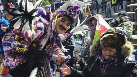 Fiona S-Hedges Photograph of Fiona pictured with one of the Mummers captains, who is dressed in a vibrant purple and gold glittery folk costume. She wears a leopard furry hat, a purple scarf and a black puffer coat. She wears small reading glasses and holds her thumb up to the camera.
