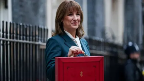 Reuters Rachel Reeves with her red Budget box standing outside 11 Downing Street, dressed in a dark green suit jacket and white blouse