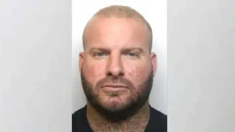 NCA Mugshot of Alex Male in a black t-shirt. He has short blond hair, a light brown beard and a neck tattoo
