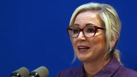 First Minister Michelle O'Neill standing in front of two microphones. She has blonde hair, tied back and she is wearing a purple jacket and dark red glasses. The background is blue. 