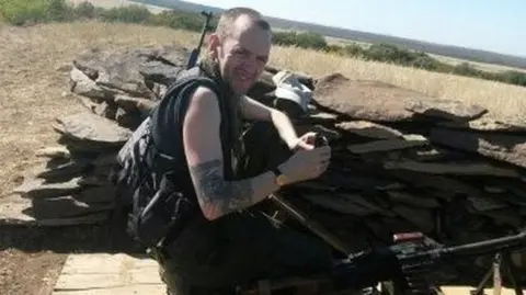 GMP A man in a vest sits in what appears to be a stone built defence. he has a shaved head and a rifle slung across his back.