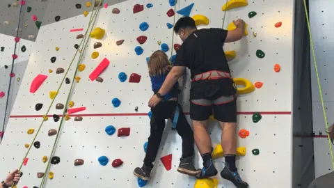 Seashell Trust Photograph of the climbing wall at Royal College Manchester. The image shows a student and a teacher climbing the wall.